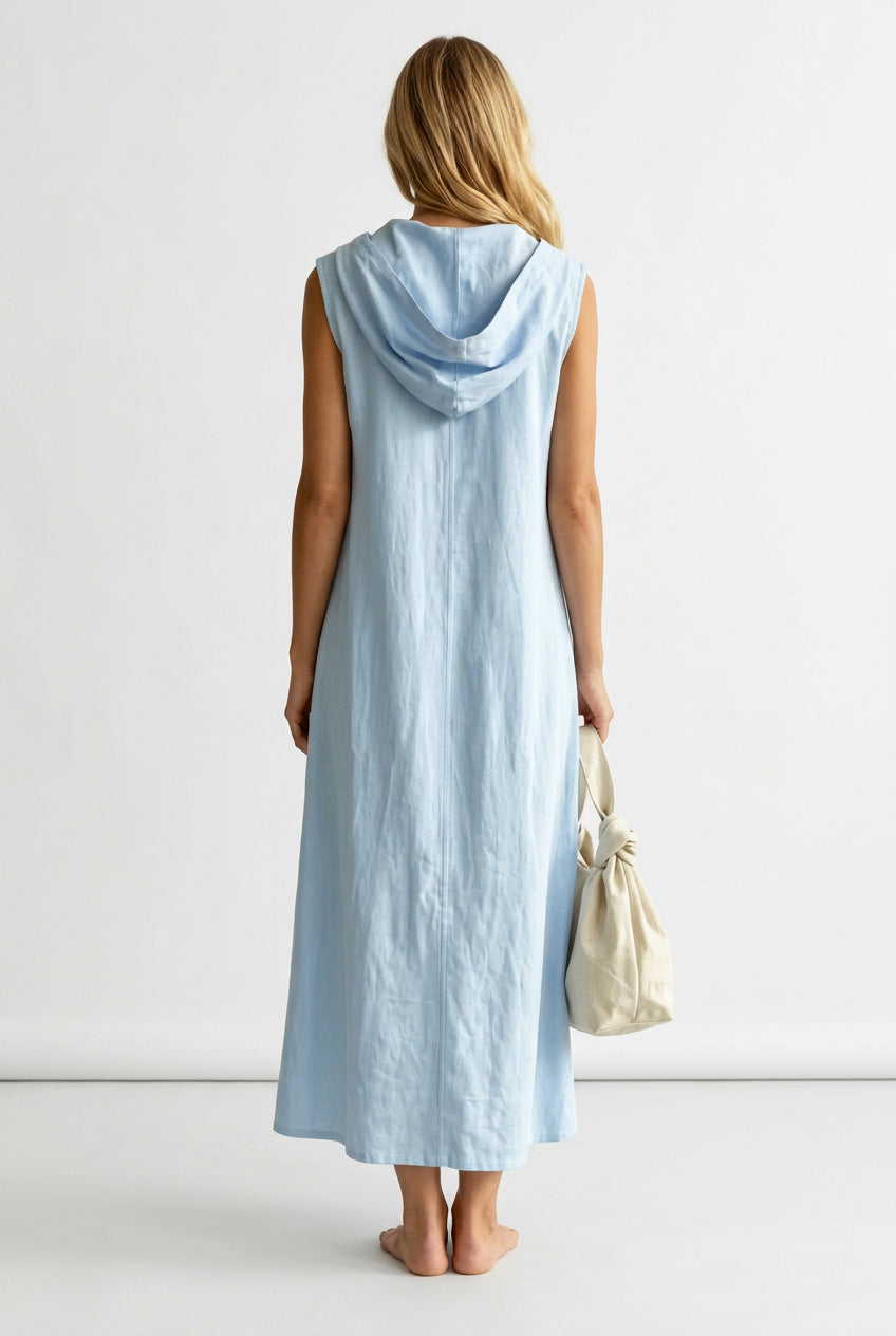 A woman viewed from behind wearing a light blue sleeveless hooded maxi dress, holding a cream-colored drawstring handbag in an outdoor garden setting with a white picket fence.