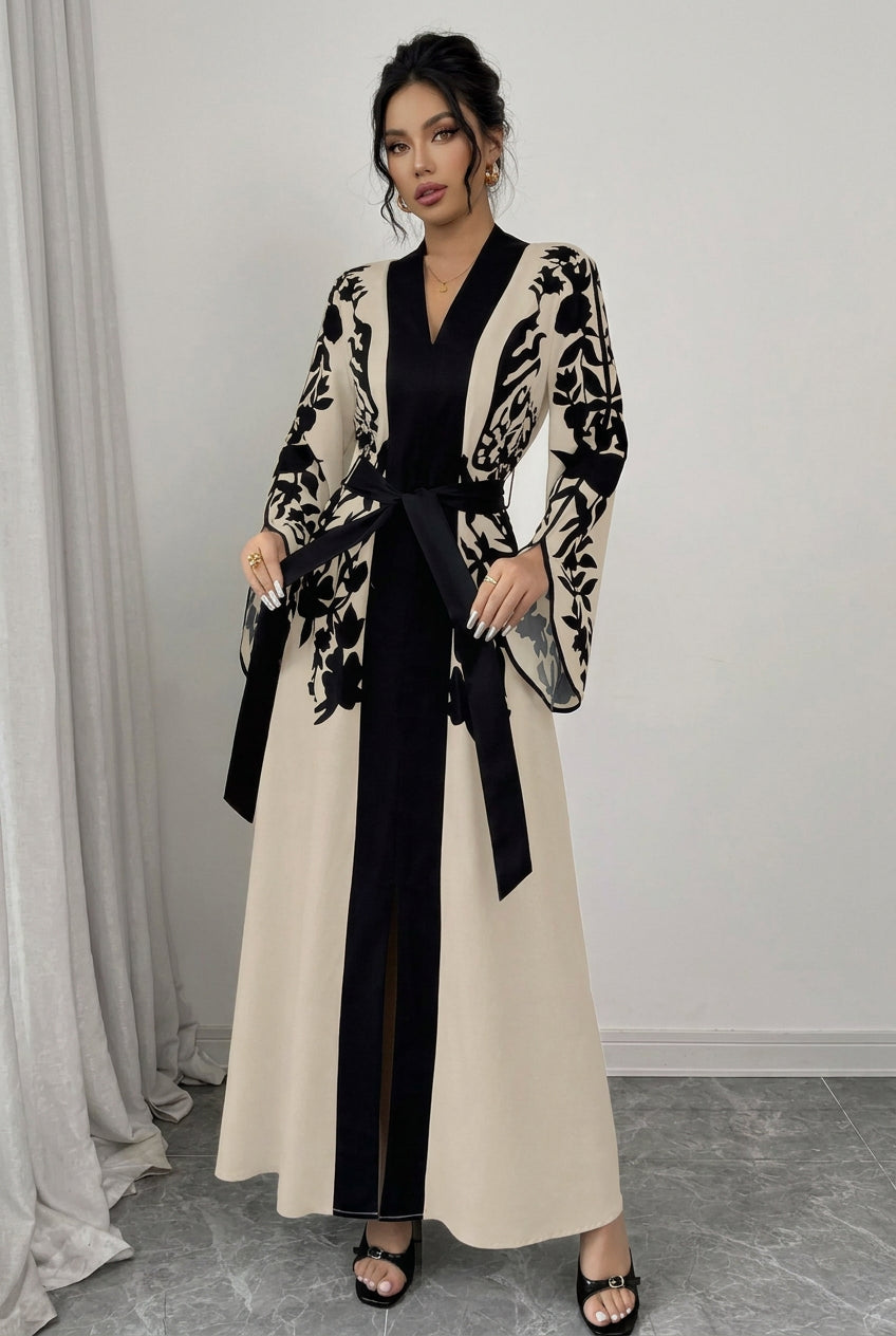 A woman models a long beige and black floral robe-style dress with a black V-neck inner layer, black waist tie, wide sleeves, and black open-toe heels against a light gray wall and gray floor.