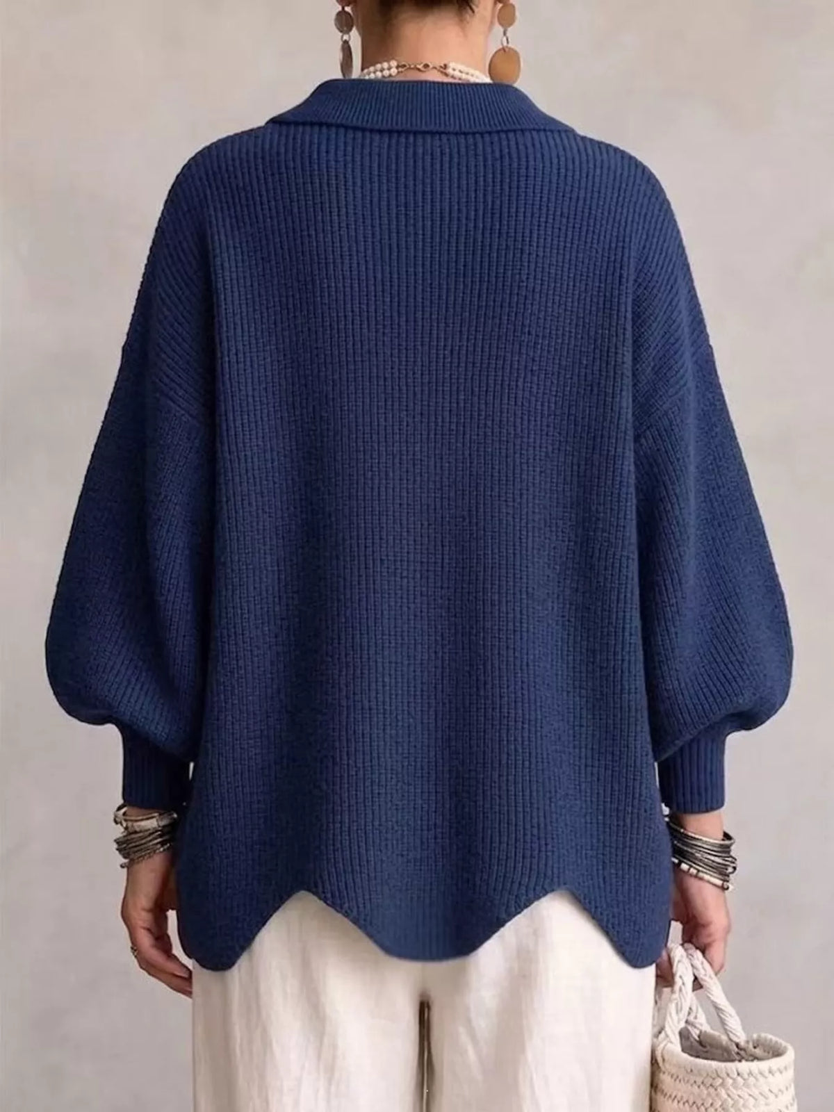 A person shown from the back wearing a navy blue ribbed knit cardigan with balloon sleeves and a scalloped hem, paired with cream pants against a neutral textured wall.
