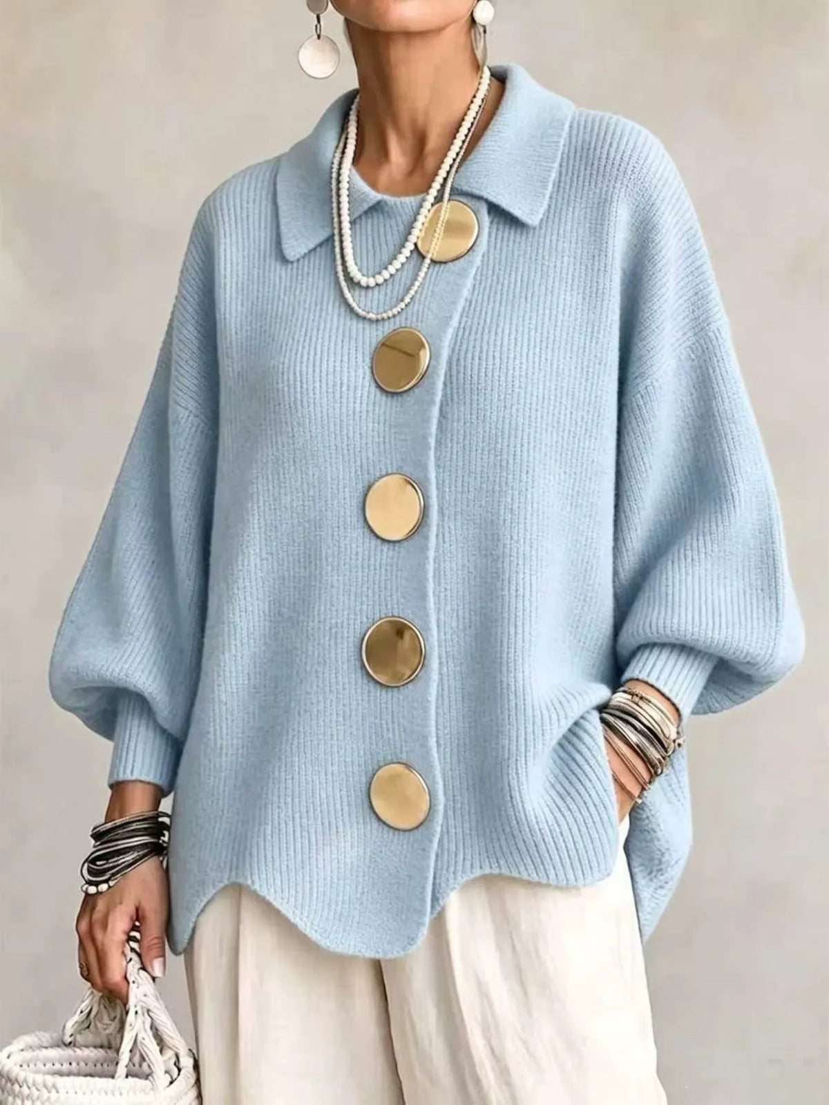 A person wearing a light blue knit cardigan with oversized round gold-tone buttons, layered pearl necklaces, bracelets, and earrings, paired with cream pants against a neutral studio background.