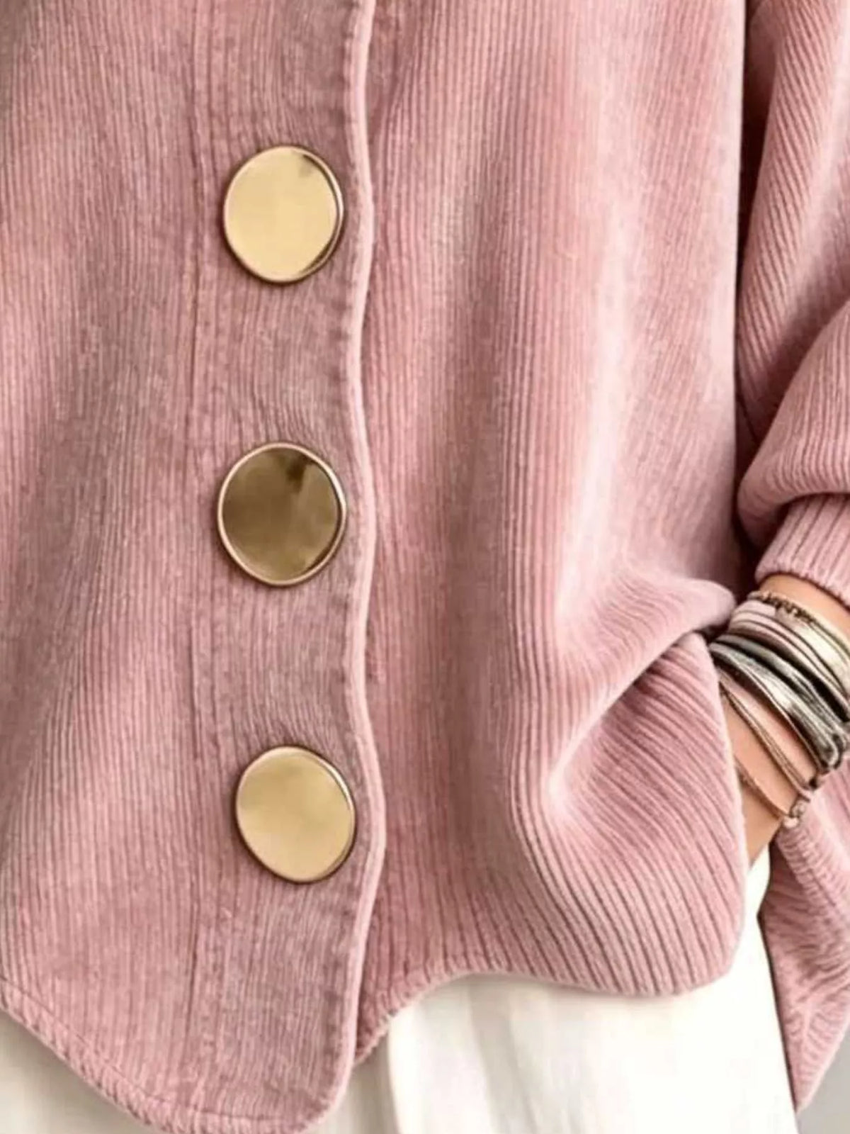Close-up of a pink ribbed corduroy cardigan with large round gold buttons, worn over a white top and accessorized with stacked silver bracelets.