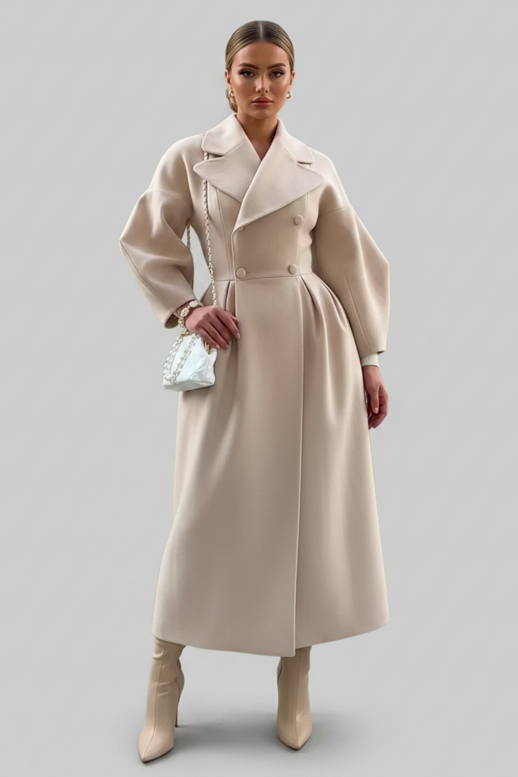 A woman wearing a long, double-breasted beige coat with wide lapels and voluminous sleeves, styled with a pearl-embellished white handbag and bracelet against a solid gray background.