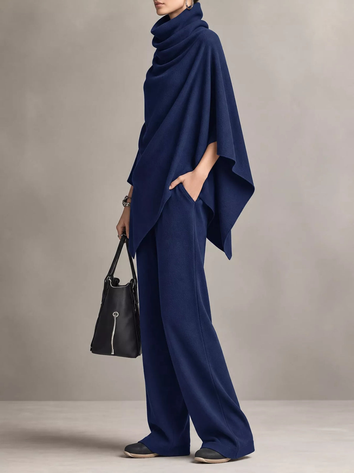 A model wearing a matching navy blue two-piece set consisting of a draped cowl-neck poncho top and wide-leg trousers, styled with a black leather handbag against a neutral gray studio background.