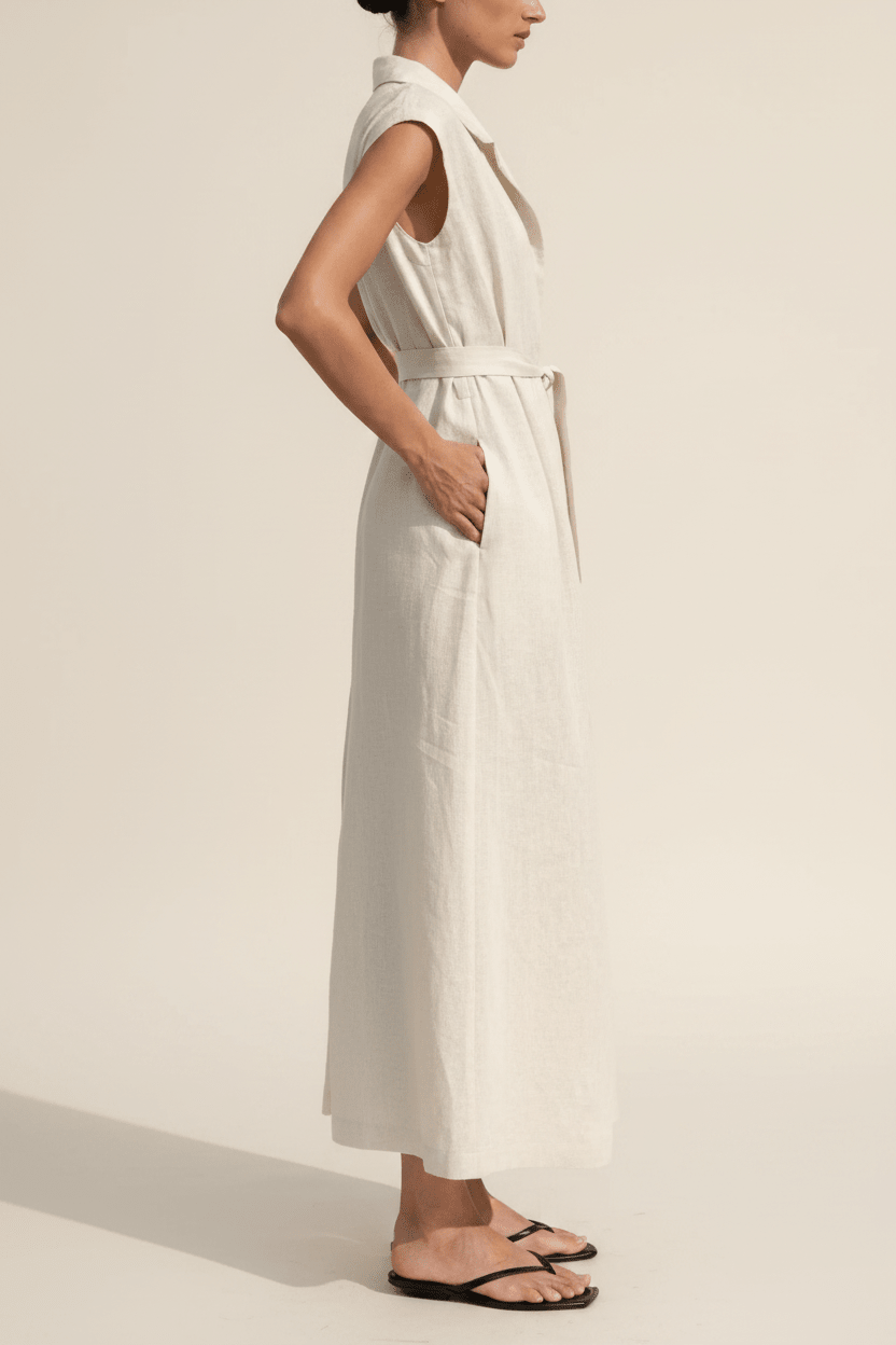 A woman wearing a sleeveless, collared, belted maxi dress in a light cream or off-white linen-like fabric, paired with black flip-flop sandals, shown in profile against a warm off-white studio background.
