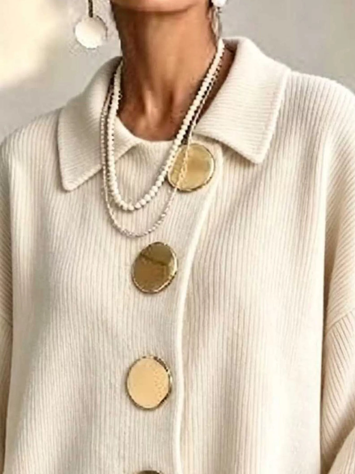 A close-up of a person wearing a cream ribbed knit collared sweater with oversized gold buttons, layered with pearl necklaces and dangling earrings.