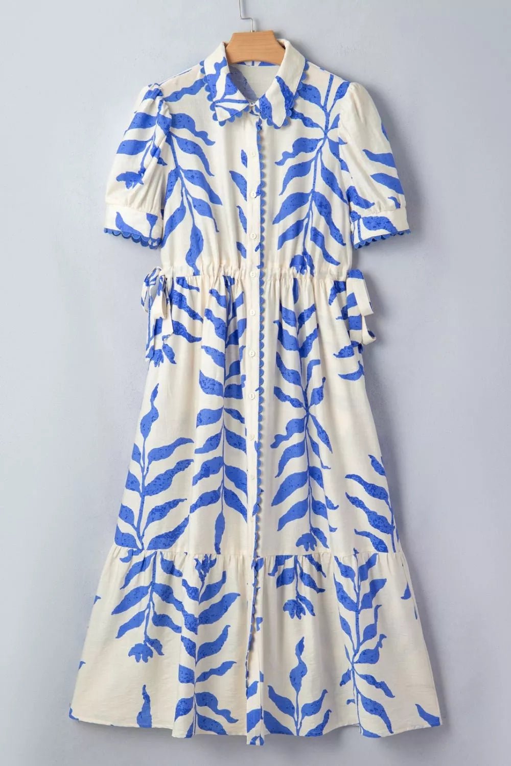 A short-sleeve collared dress with a blue botanical leaf print on a cream background, hanging on a wooden hanger against a light gray wall.