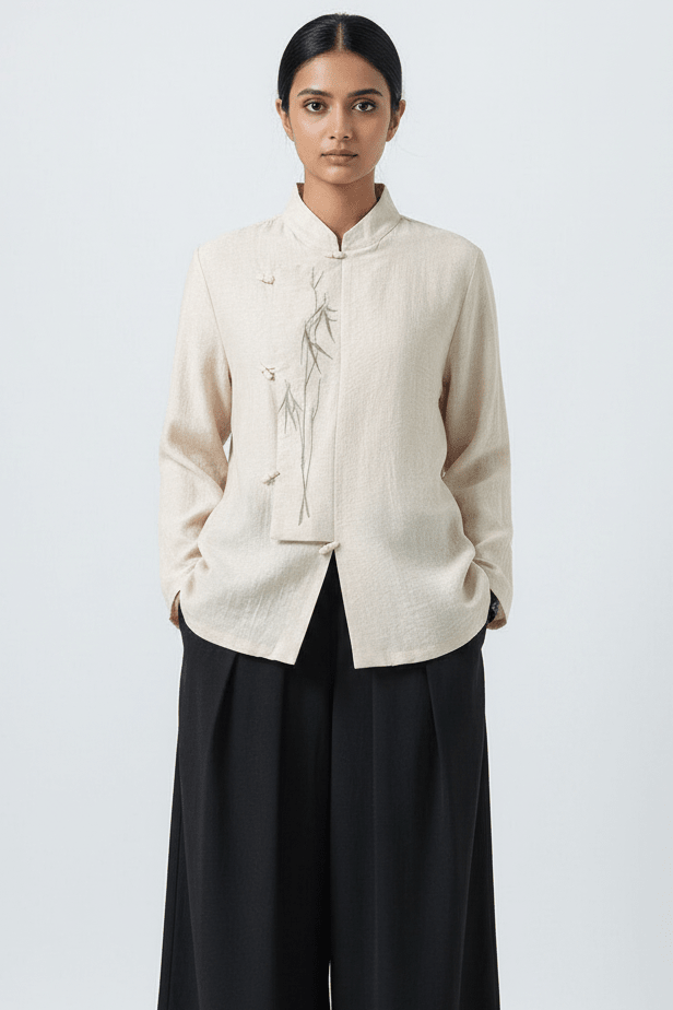 A woman modeling a light beige linen long-sleeve top with a mandarin collar and delicate botanical embroidery on the front, paired with black wide-leg trousers, against a light gray studio background.