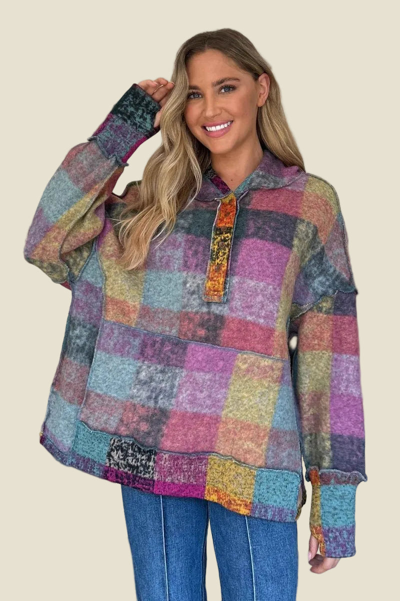 A woman wearing a multicolor patchwork plaid pullover hoodie with a half-placket and ribbed cuffs, paired with blue jeans, against a solid beige background.