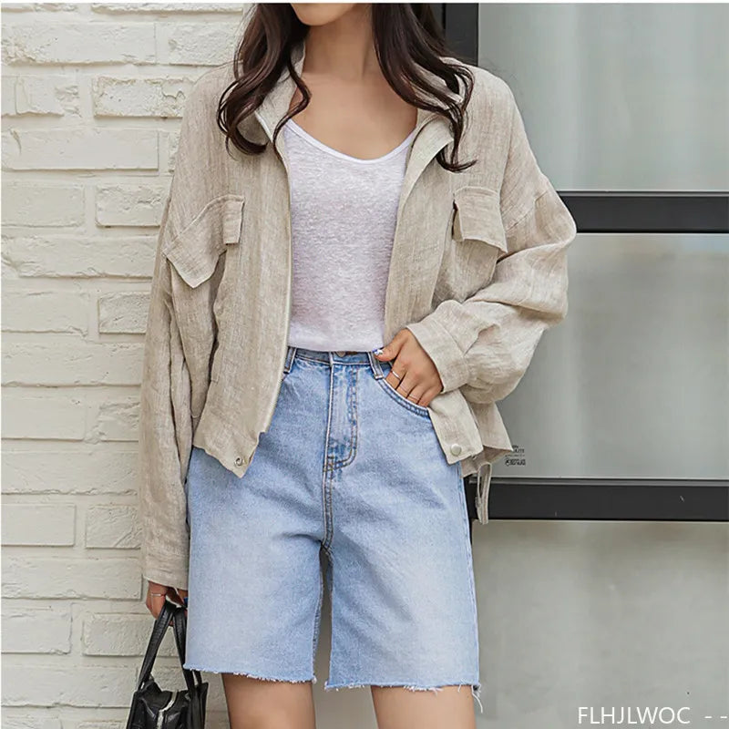 Person wearing a beige jacket, white shirt, and denim shorts against a stone wall.
