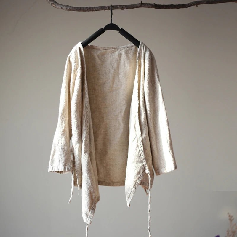 A light beige linen open-front cardigan with long sleeves and drawstring ties, hanging on a black hanger from a rustic wooden branch against a neutral beige wall.