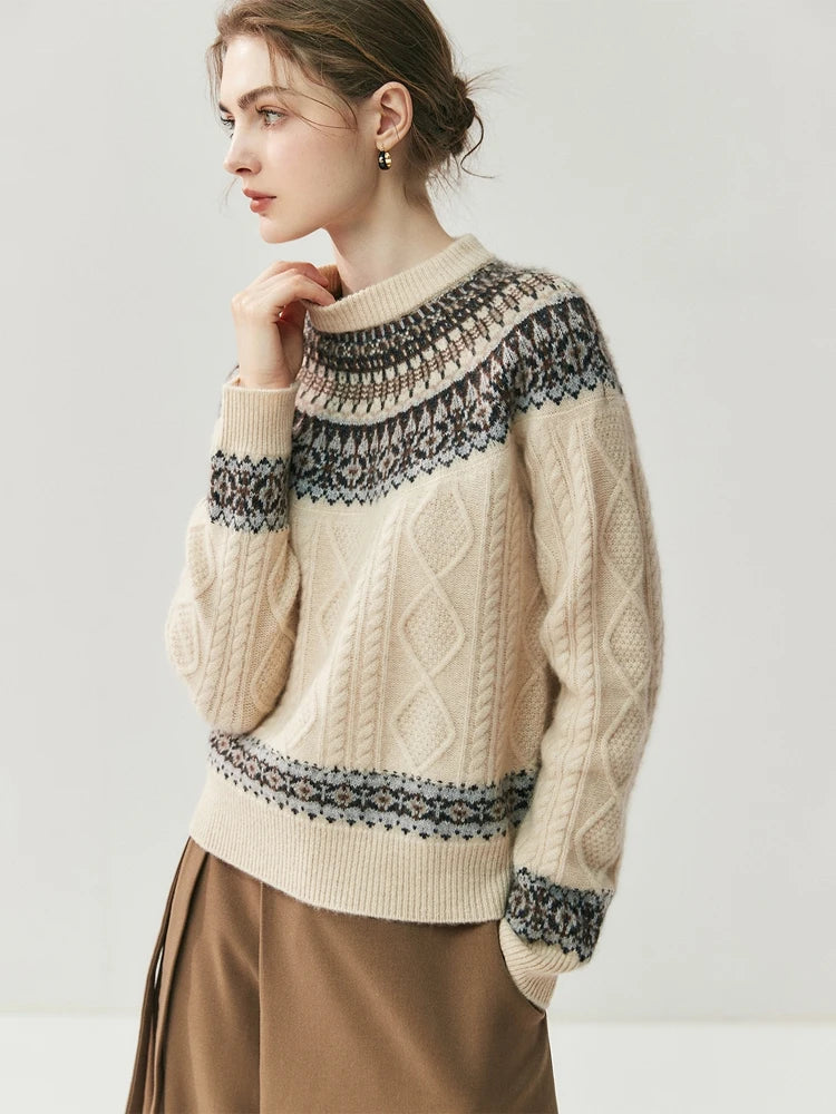 A woman wearing a cream-colored cable-knit sweater with dark gray and navy Fair Isle patterns across the yoke, cuffs, and hem, paired with brown pleated trousers, set against a plain light gray studio background.
