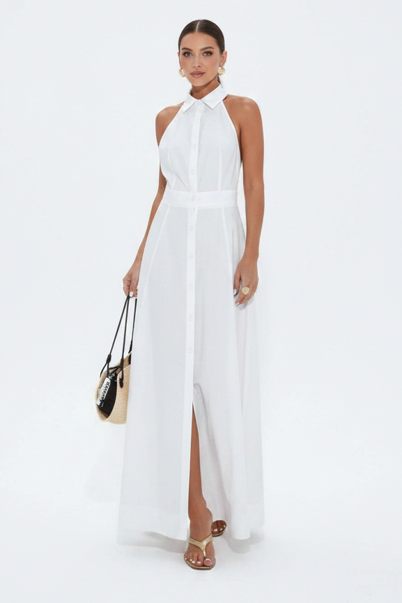 A woman modeling a white sleeveless halter-neck maxi dress with a pointed collar, front button placket, and high front slit, standing against a plain white studio background and holding a small black-and-beige handbag.