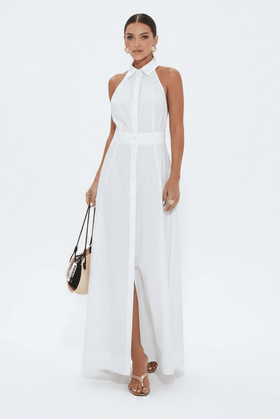 A woman modeling a sleeveless white halter-neck maxi dress with a high front slit, holding a small beige and black handbag against a plain white studio background.