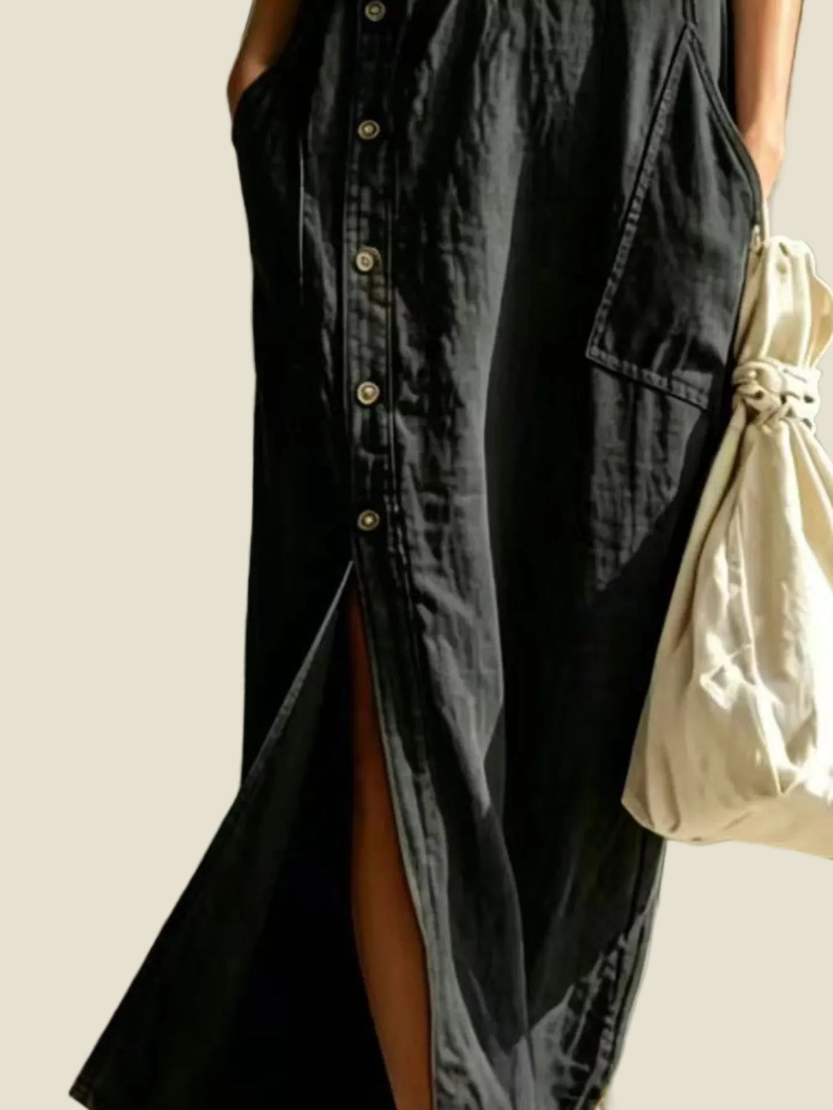 A close-up of a person wearing a black button-front midi dress with a high side slit, holding a cream drawstring tote bag outdoors.
