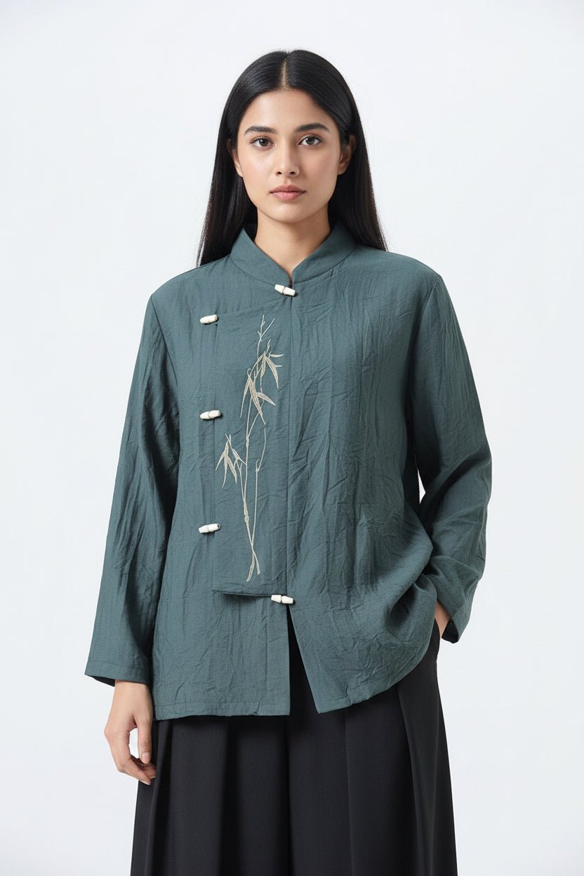 A woman modeling a dark teal-green long-sleeve jacket with a textured crinkled fabric, featuring a bamboo plant embroidery on the front and white wooden toggle closures, paired with a black skirt against a light gray background.