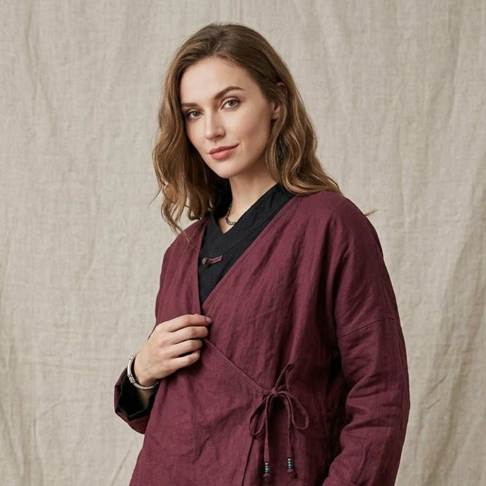 A woman wearing a burgundy wrap-style linen jacket with a drawstring waist over a black top and black skirt, standing against a beige linen backdrop.