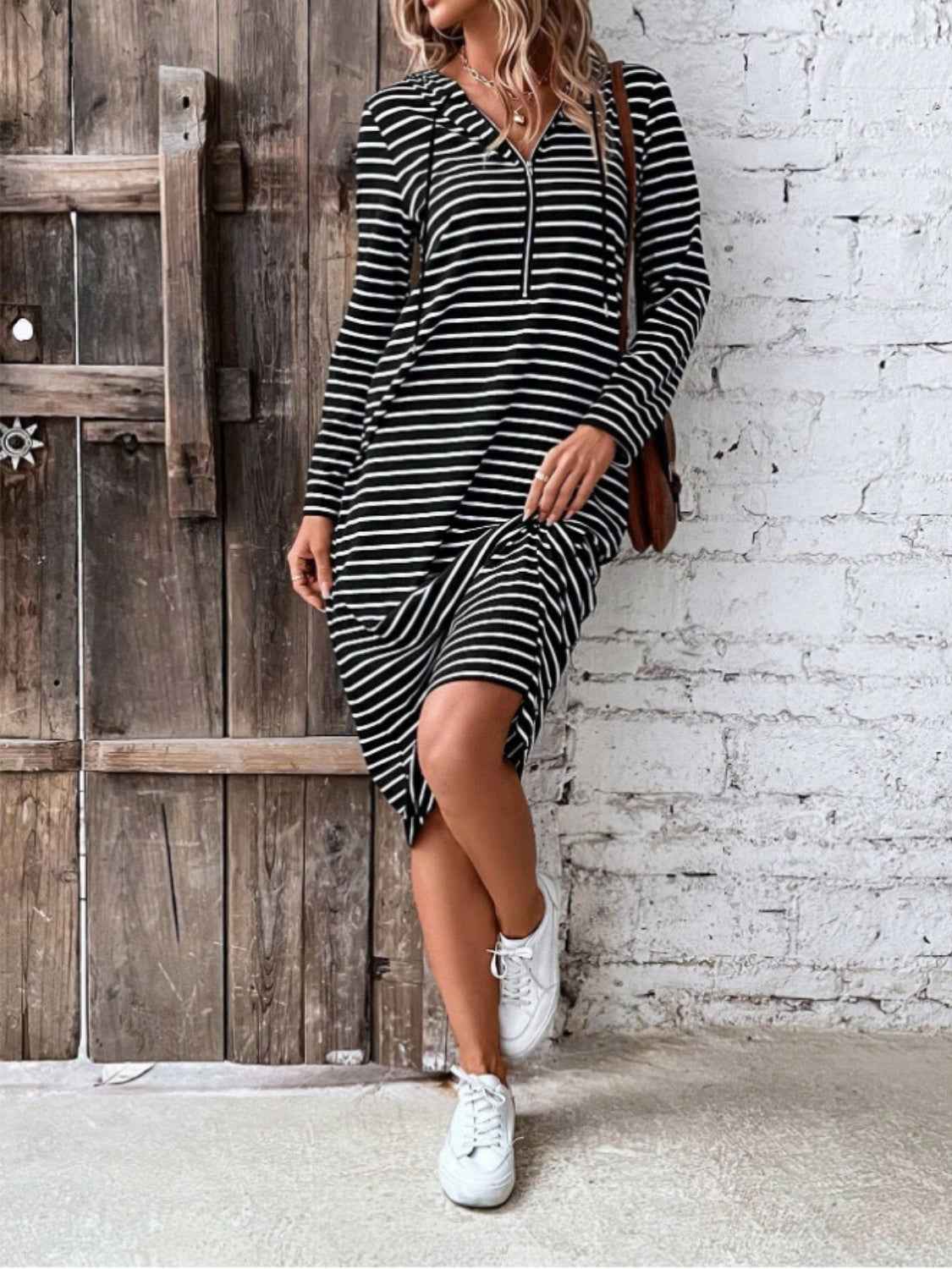 Woman wearing a black and white striped dress with white sneakers in front of a wooden door and white brick wall.