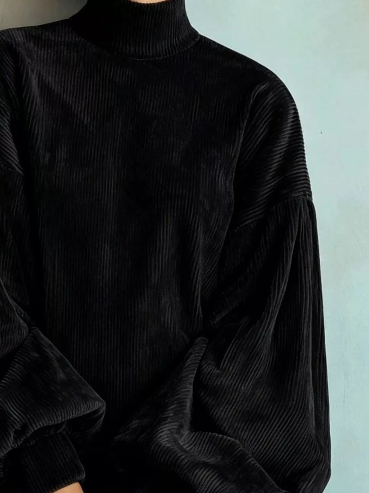A close-up of a person wearing a black ribbed corduroy mock-neck sweater with voluminous balloon sleeves, set against a pale blue wall.