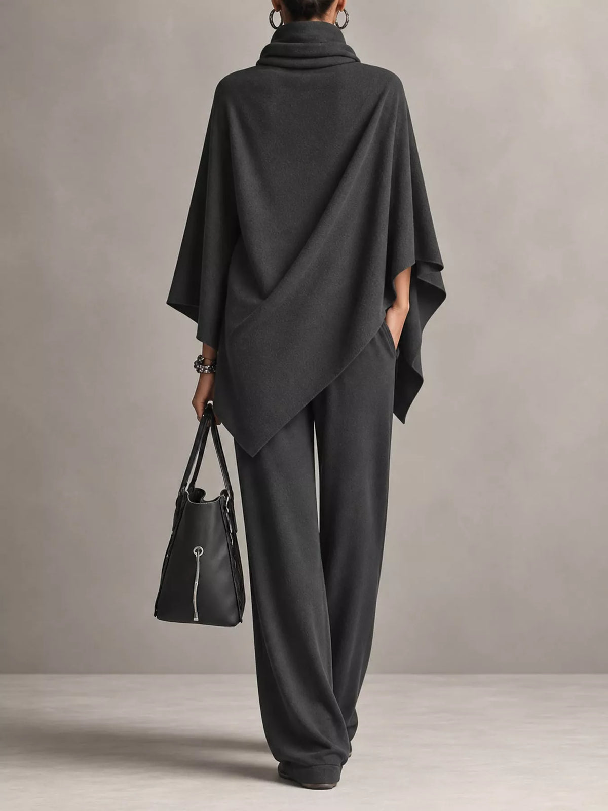 A model from behind wearing a dark charcoal gray cowl-neck poncho and matching wide-leg trousers, holding a black leather handbag against a neutral gray studio background.