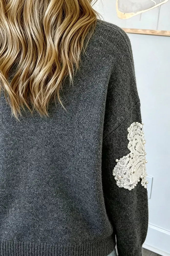 A woman viewed from behind wearing a dark gray knit sweater with a white floral lace applique on the sleeve, paired with light blue denim shorts.