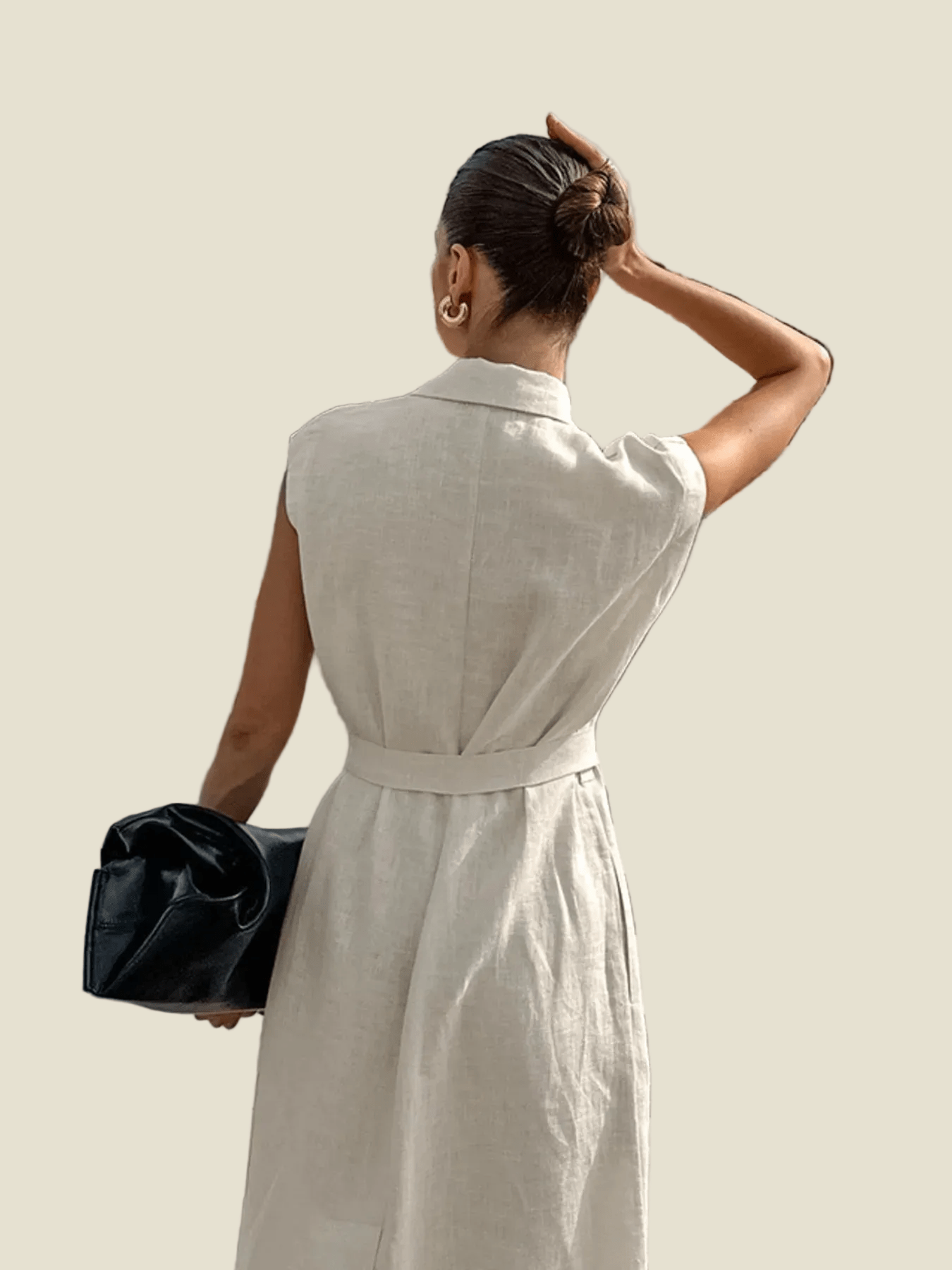 A woman shown from behind wearing a light beige linen belted dress, holding a black clutch bag against a solid beige background.
