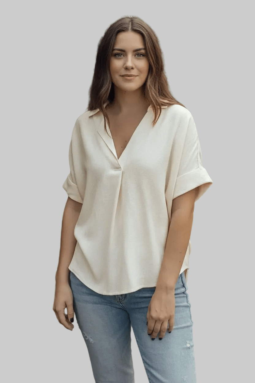 A woman modeling a cream-colored short-sleeve blouse with a V-neckline and light-wash blue jeans, set against a solid gray background.