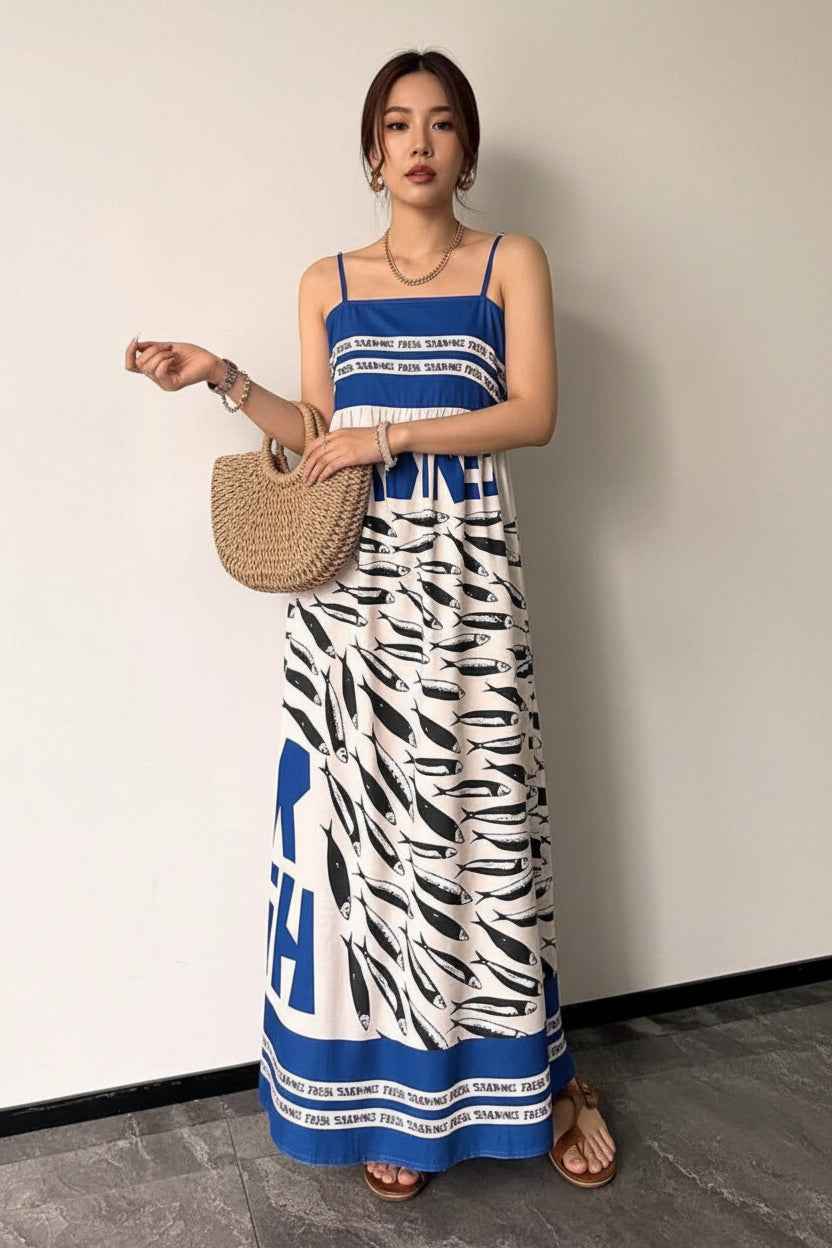 Woman wearing a blue and white patterned dress holding a straw bag against a plain background