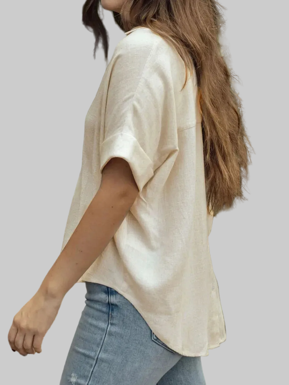A woman wearing a loose, short-sleeve cream-colored top and light blue jeans, shown from a side-back angle against a solid gray background.
