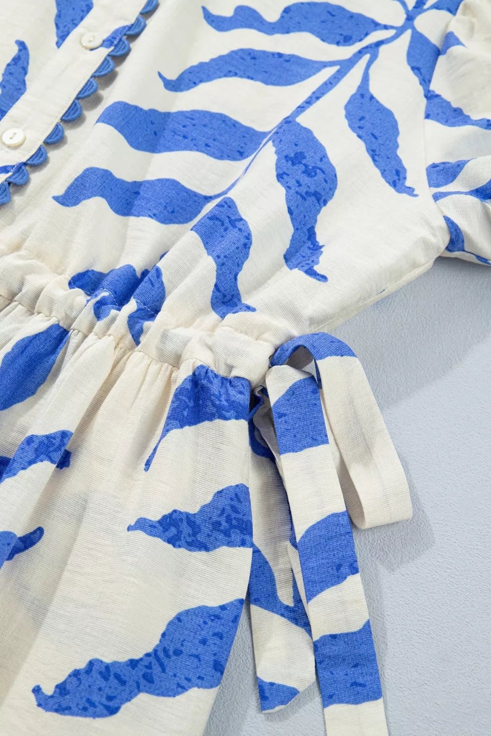 A close-up of a white fabric garment with a bold blue abstract leaf-like print, featuring a gathered waist and matching tie detail.