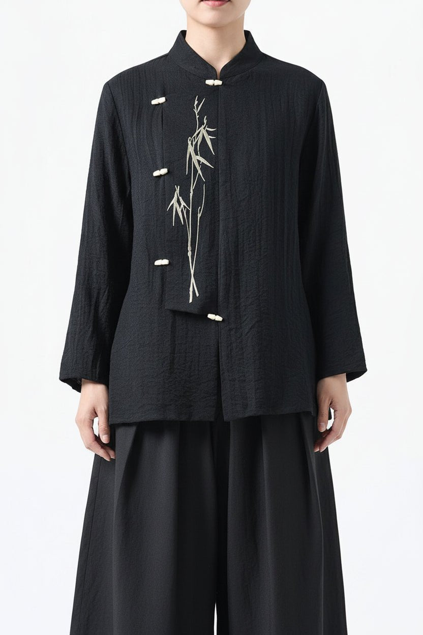 A person wearing a black long-sleeve cropped jacket with a mandarin collar, white decorative buttons, and a white bamboo plant embroidery on the front, paired with black wide-leg pants, against a plain white background.