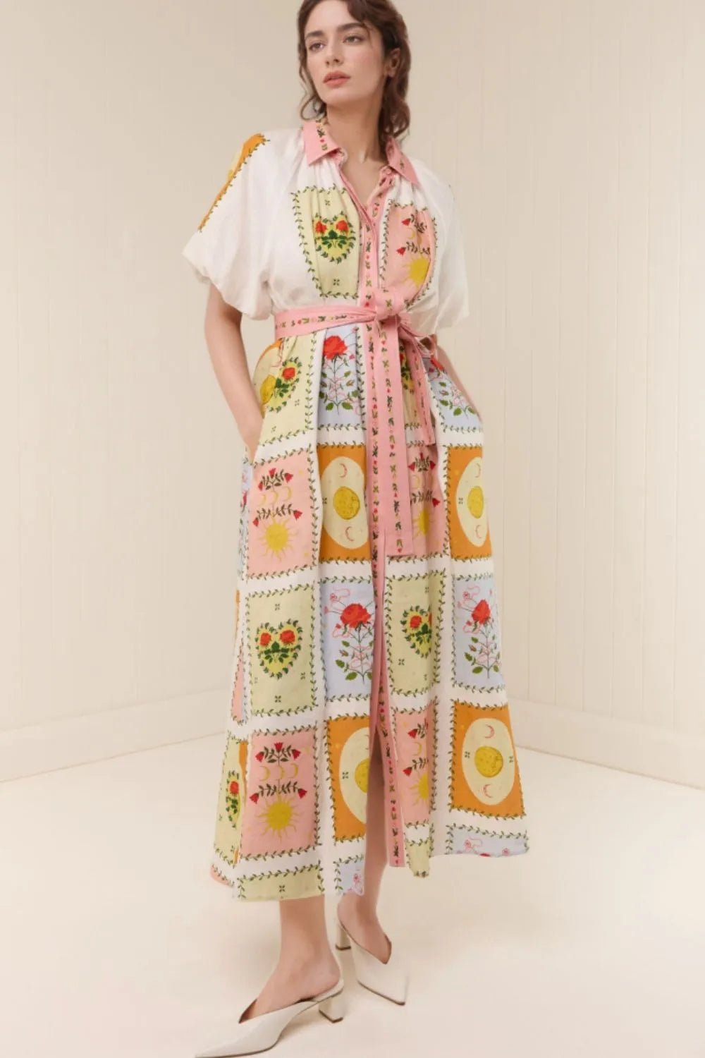 A woman models a long white shirt dress with a colorful patchwork print featuring floral and egg motifs, styled with a pink waist tie and white pointed-toe heels against a cream-colored studio background.