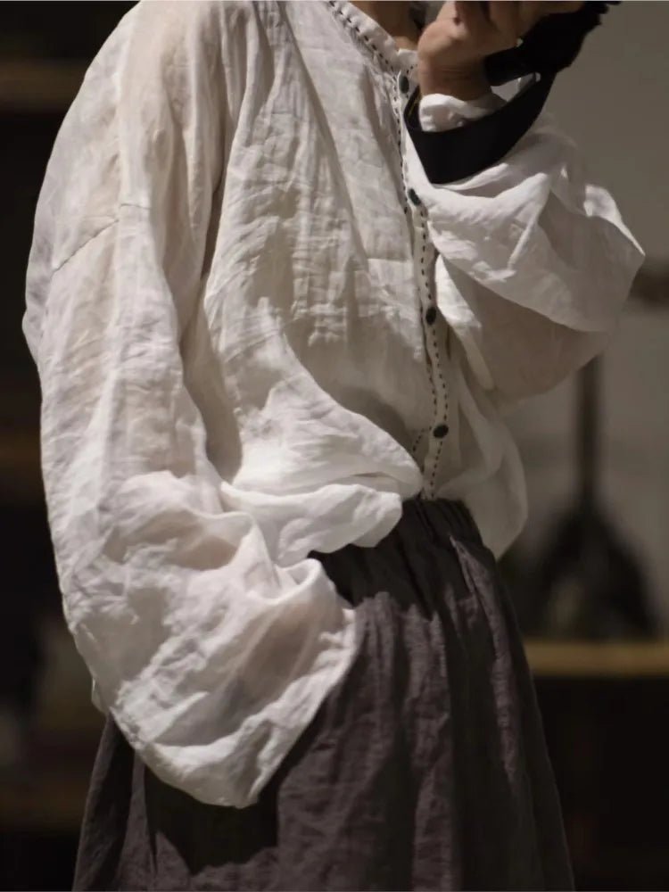 A person wearing a loose white long-sleeve blouse with gathered cuffs and a button placket, paired with a dark skirt, photographed in a dim indoor setting.