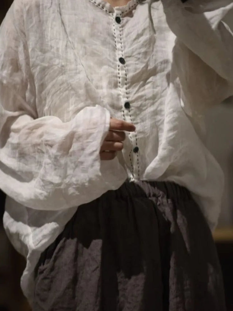 A person wearing a loose white long-sleeve blouse with a decorative beaded neckline and dark buttons, paired with a dark skirt, photographed in a dim indoor setting.