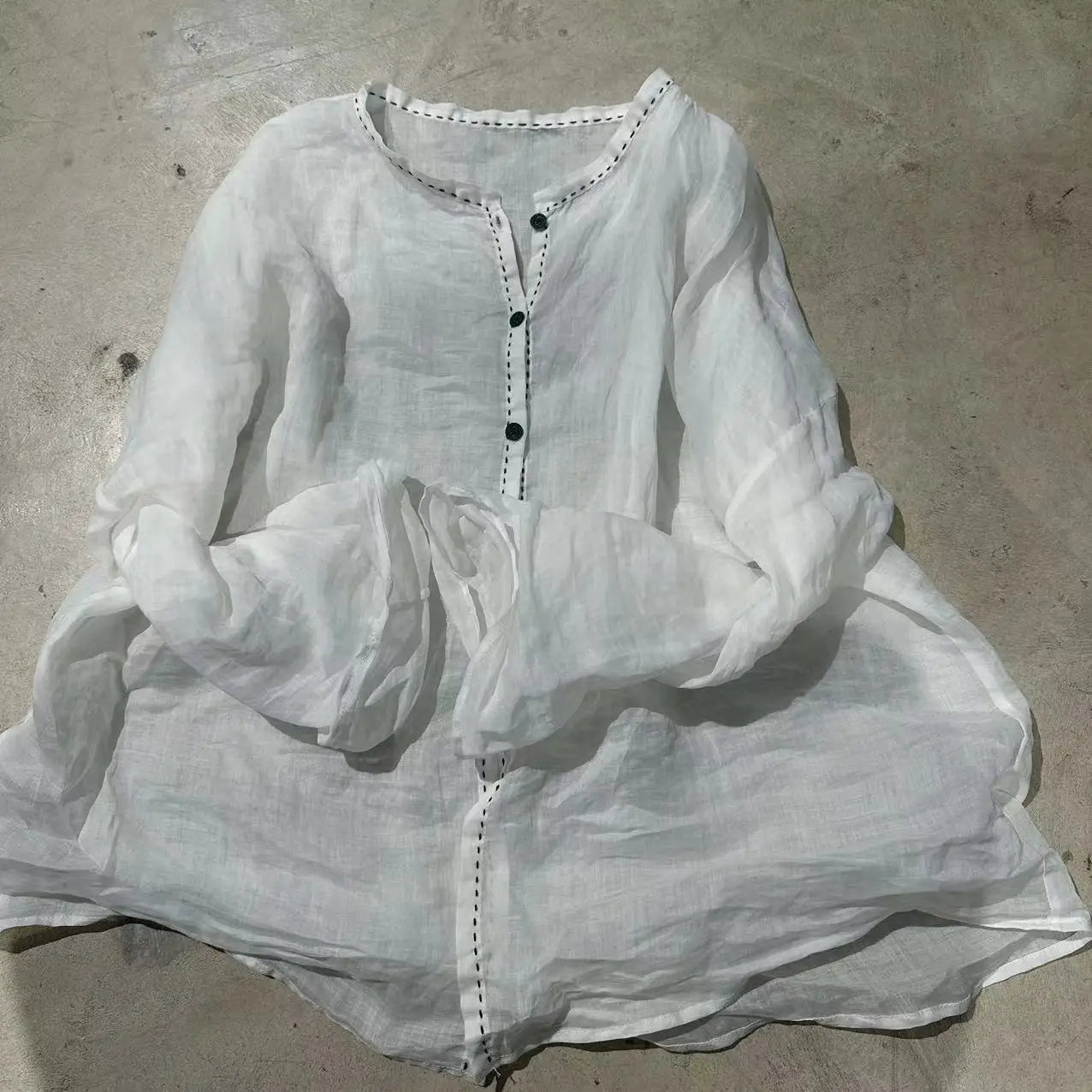 A white long-sleeve linen tunic top with black buttons and decorative stitching, laid flat on a concrete floor.