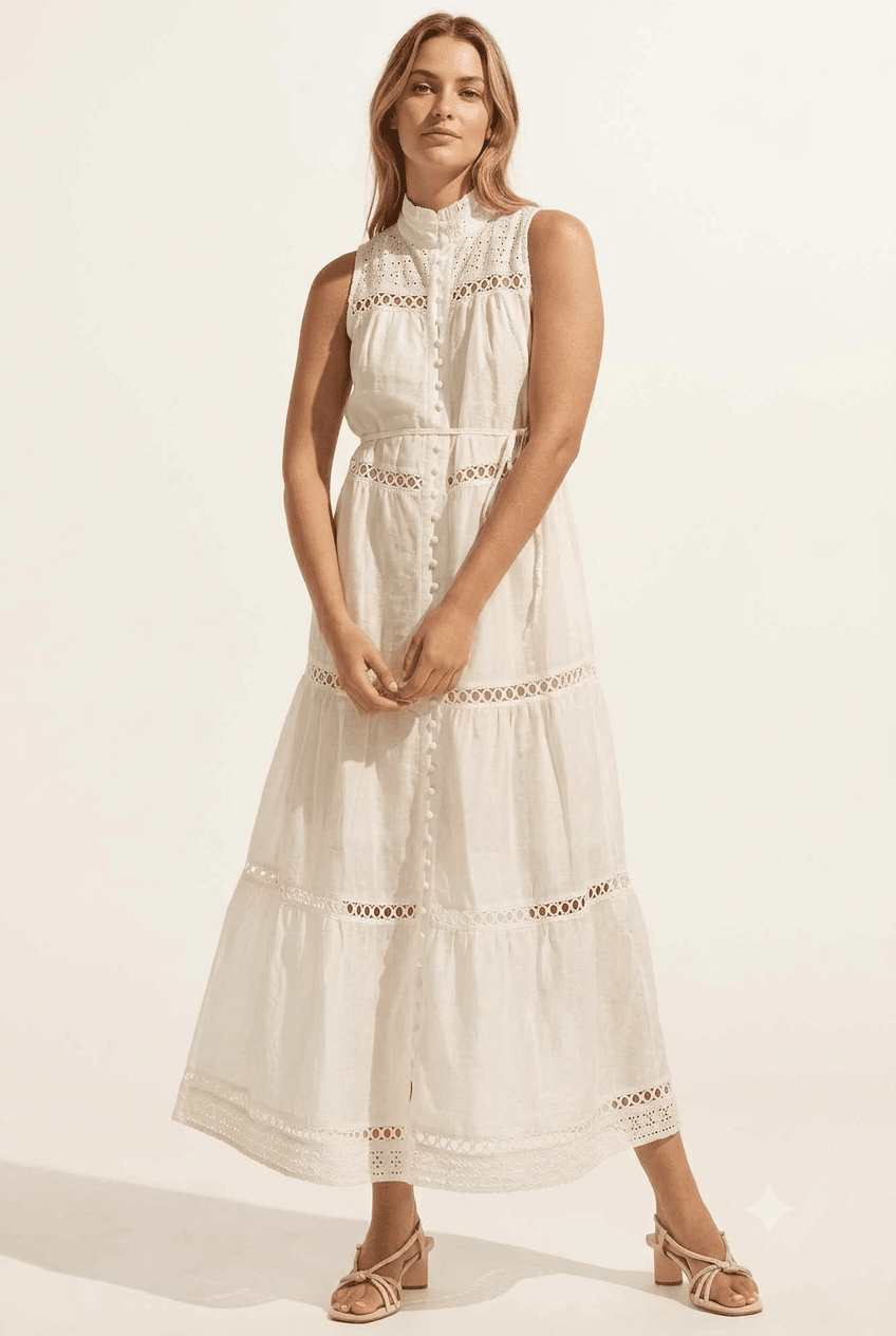A woman modeling a sleeveless, high-neck, tiered white maxi dress with intricate eyelet embroidery, paired with beige strappy sandals, set against a plain off-white studio background.