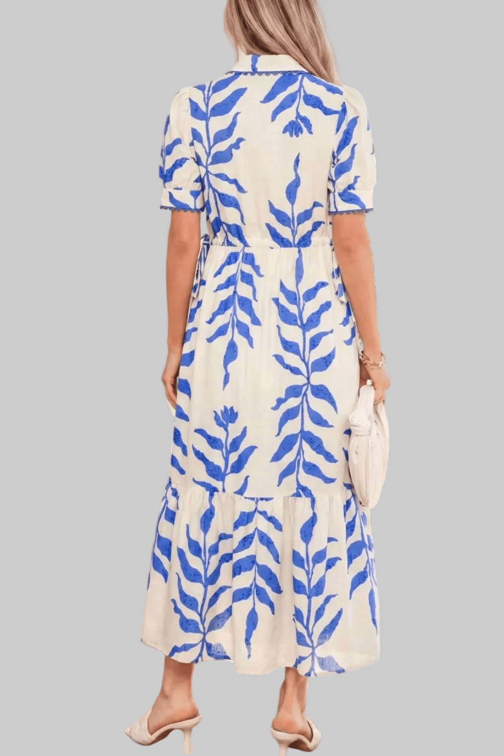 A woman shown from the back wearing a short-sleeve midi dress with a blue leaf pattern on a cream background, holding a cream clutch against a solid gray background.