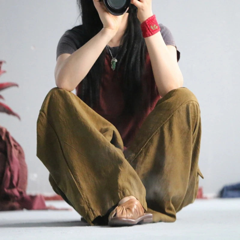 A person sitting cross-legged on a light floor, holding a camera to their face. They are wearing loose, wide-leg olive brown pants, a dark gray t-shirt, and brown slip-on shoes, with a red patterned wristband and a green pendant necklace.