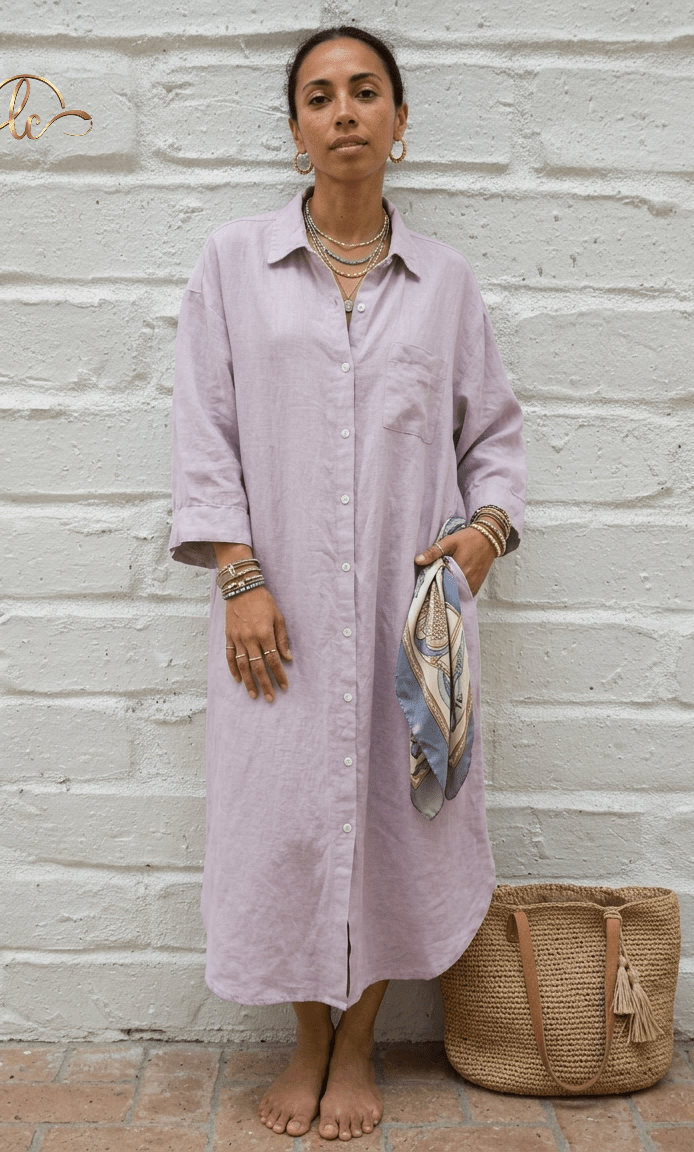 A woman wearing a light purple shirt dress standing against a white brick wall with a woven bag beside her.