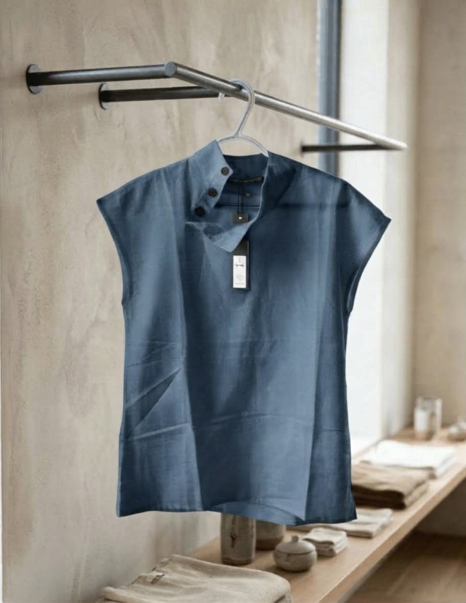 A sleeveless dusty blue linen shirt with a mock neck and three buttons, hanging on a metal hanger from a clothing rack in a rustic, warm-toned room.