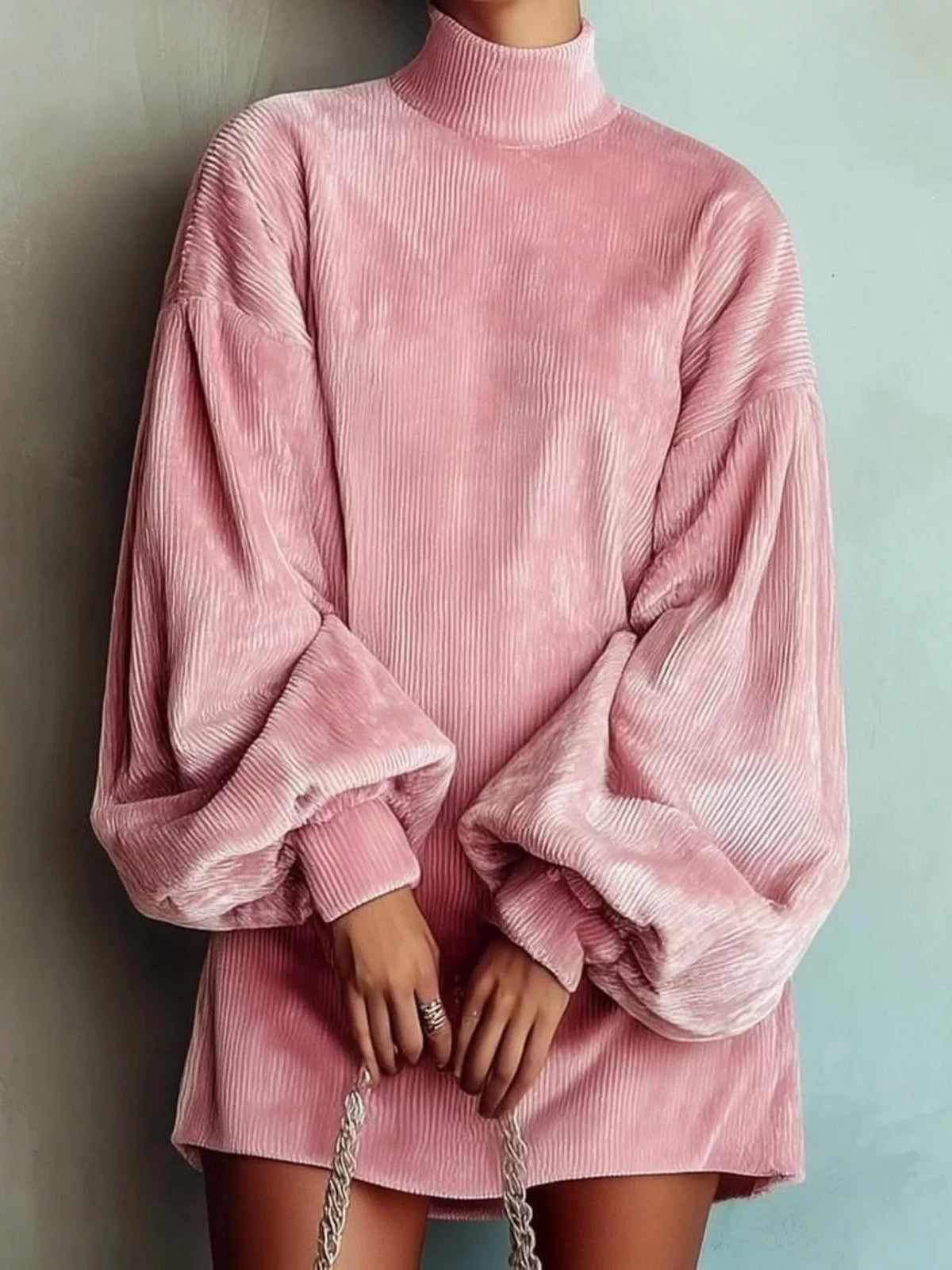 A person wearing an oversized pink corduroy mock-neck tunic dress with voluminous balloon sleeves, standing against a pale blue wall and holding a silver chain handbag.