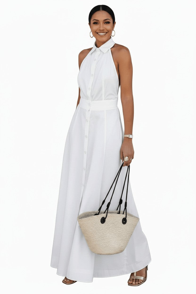 A woman modeling a sleeveless white halter-neck maxi dress with a button-front bodice and a matching waistband, holding a woven straw tote bag against a plain white studio background.