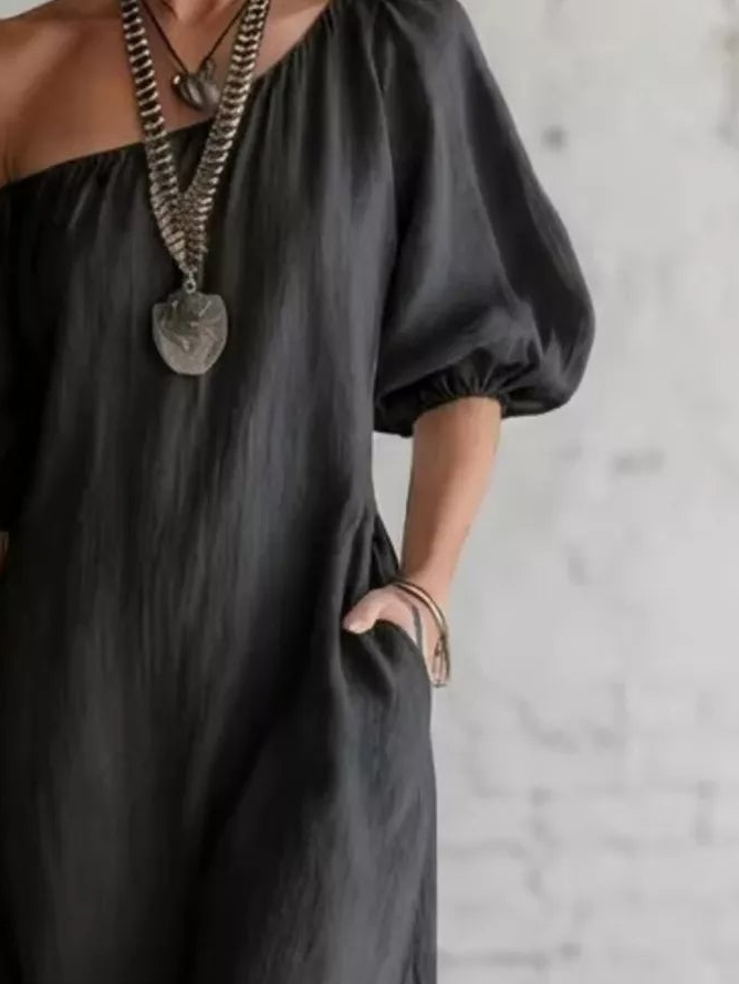 A close-up of a person wearing a loose, one-shoulder black dress with puff sleeves and side pockets, styled with layered silver chain necklaces and bracelets against a light textured wall.