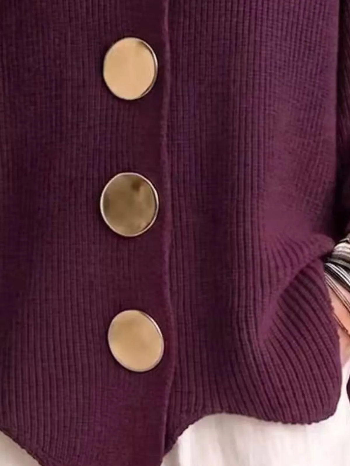 Close-up of a ribbed knit cardigan in deep plum purple with three large round metallic buttons, shown on a person wearing a white top.