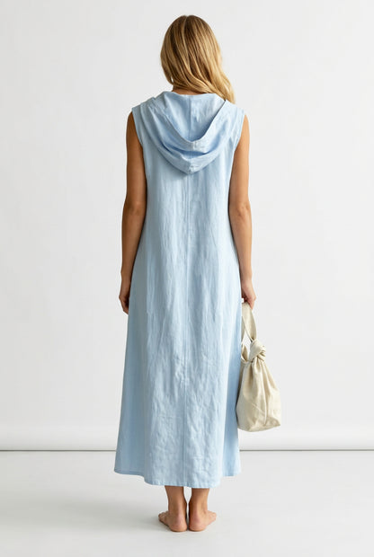 A woman viewed from behind wearing a light blue sleeveless hooded maxi dress, holding a cream-colored drawstring handbag in an outdoor garden setting with a white picket fence.