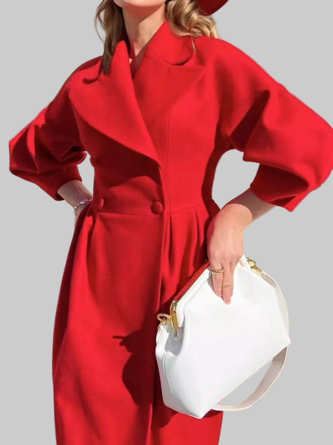 A woman wearing a bright red double-breasted coat with balloon sleeves and holding a white crescent-shaped handbag with gold hardware, set against a solid gray background.