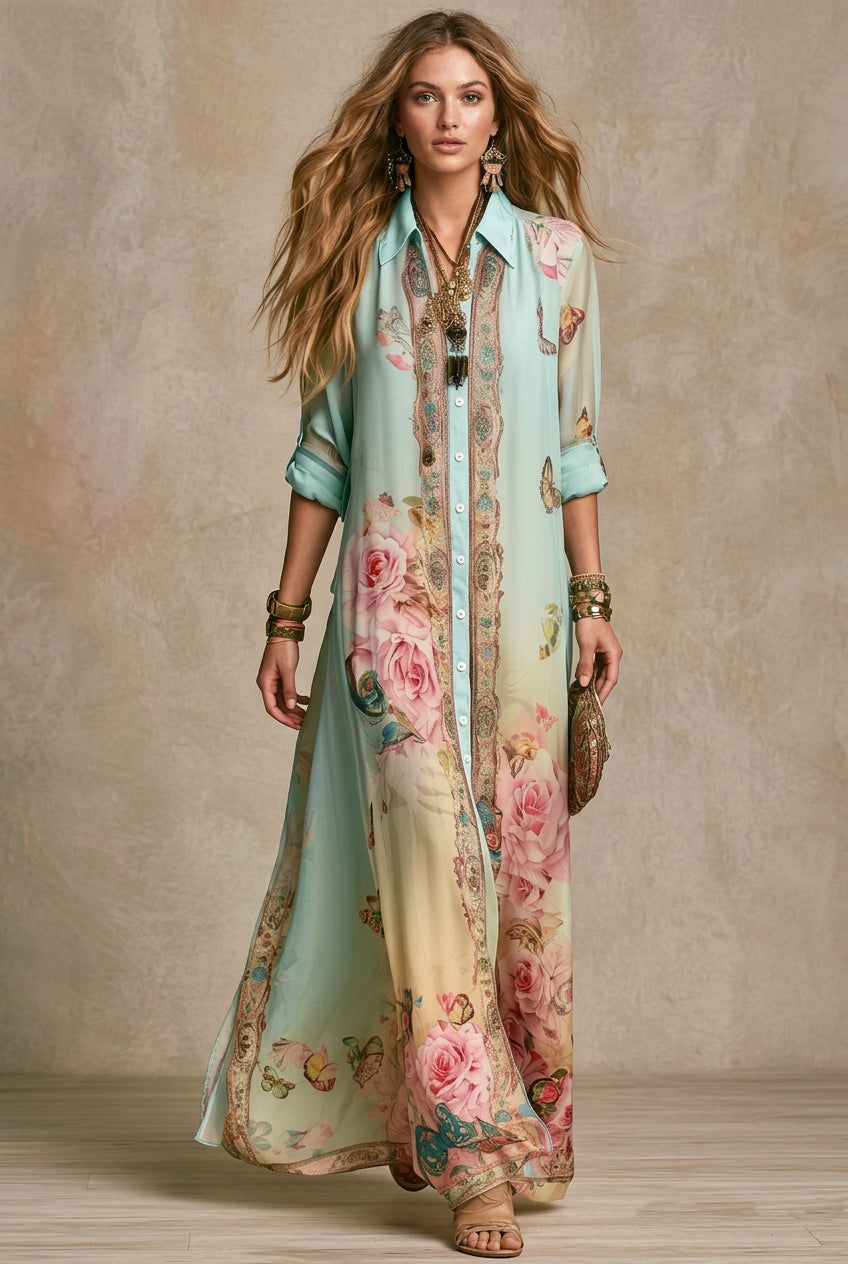 A woman models a long, flowing button-up maxi dress with a light blue and cream gradient, decorated with pink rose and butterfly floral prints and ornate gold borders. She is accessorized with gold jewelry and a small brown clutch, standing against a textured beige studio backdrop.
