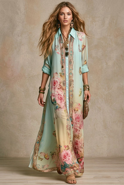 A woman models a long, flowing button-up maxi dress with a light blue and cream gradient, decorated with pink rose and butterfly floral prints and ornate gold borders. She is accessorized with gold jewelry and a small brown clutch, standing against a textured beige studio backdrop.