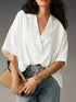 Person wearing a white blouse and holding a beige handbag against a gray background