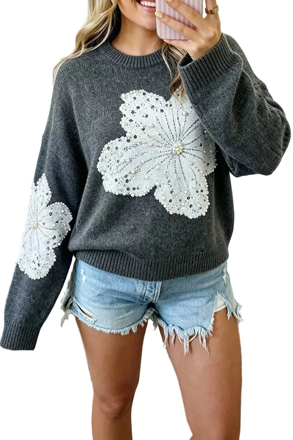 A woman wearing a dark gray knit sweater with large white floral lace appliques on the front and sleeves, paired with light blue denim shorts, shown in a mirror selfie.