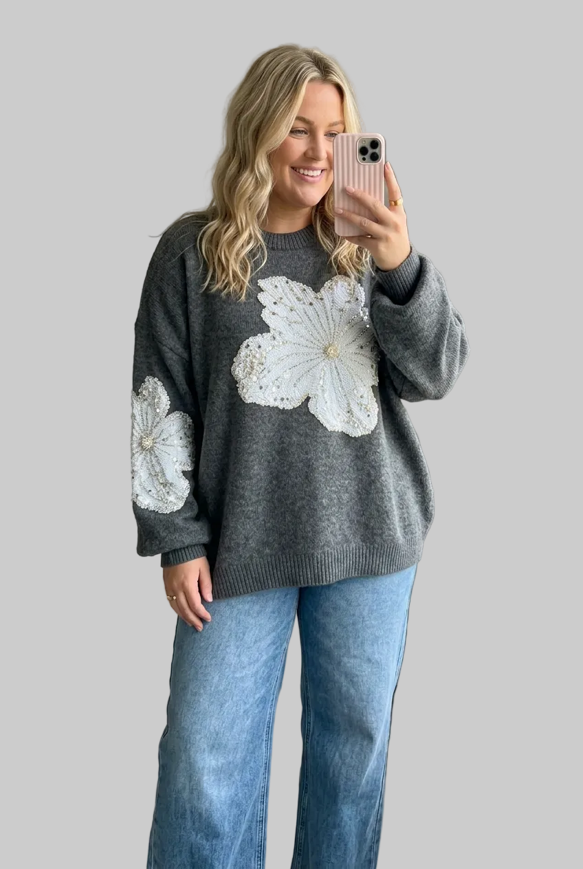 A woman takes a mirror selfie wearing a dark gray knit sweater with large white crochet floral details on the front and sleeve, paired with light blue jeans.
